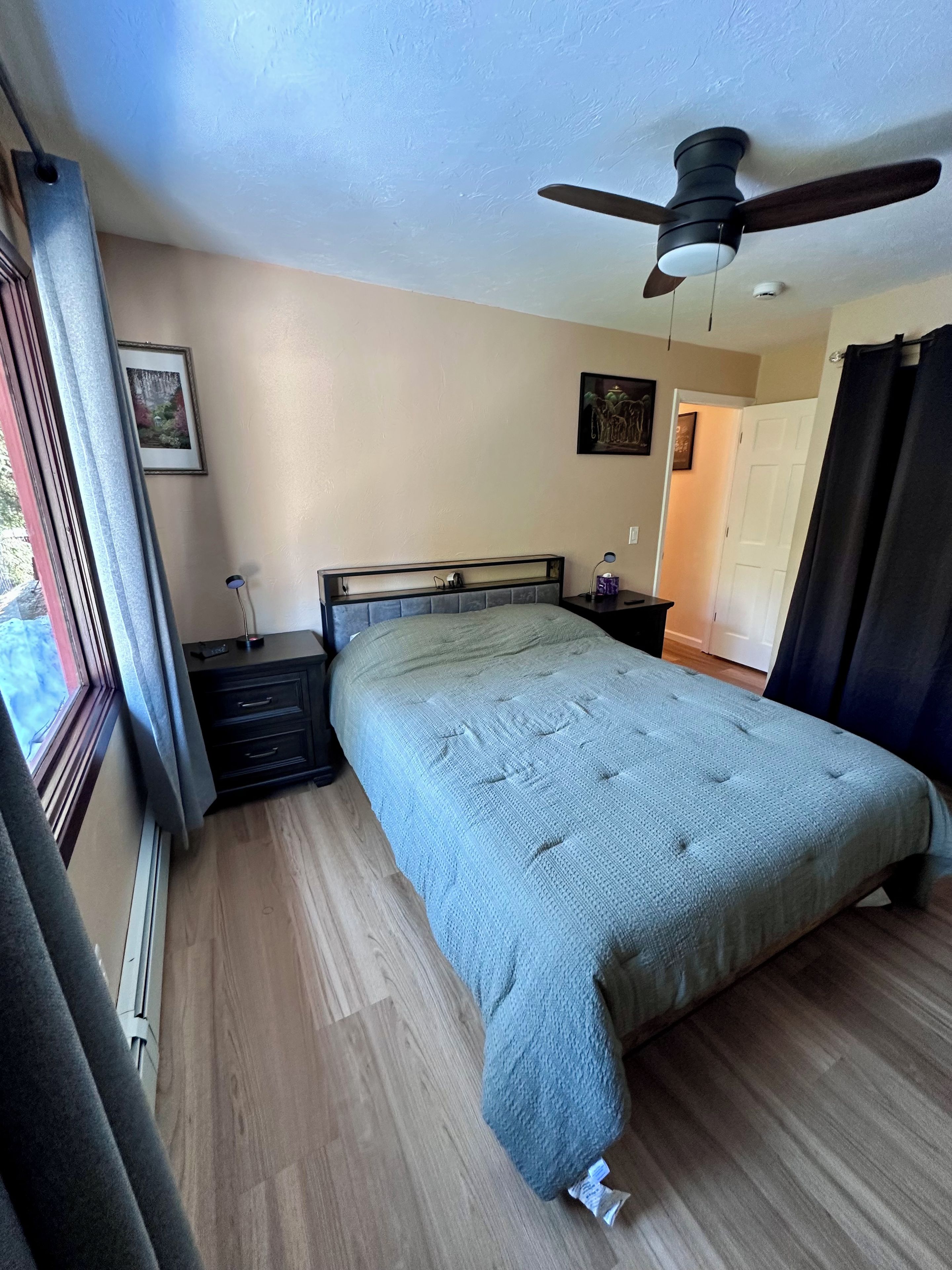 Une chambre à coucher comprenant un lit, une table de chevet, un ventilateur de plafond, une fenêtre avec des rideaux et une porte.