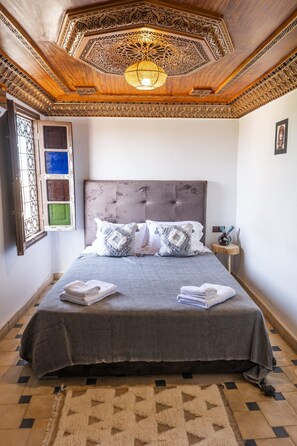 A bedroom with a large bed, a decorative ceiling, and a window with colored glass panes. - Charming 3-bedroom riad in Marrakech with cool AC breeze (Marrakech)
