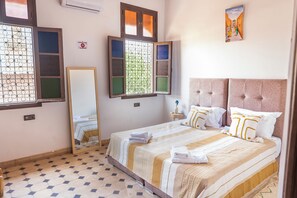 A hotel room with a bed, a mirror, a window with multiple panes, a small bedside table with a vase, and a wall-mounted artwork. - Charming 3-bedroom riad in Marrakech with cool AC breeze (Marrakech)
