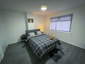 Ein Schlafzimmer mit einem Bett, Nachttisch, Lampe und einem Fenster mit Jalousien.