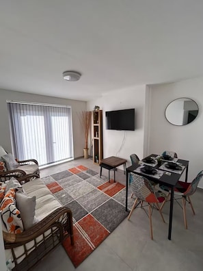 Ein Wohnzimmer mit einer Couch, einem Esstisch mit Stühlen, einem Flachbildfernseher und einem großen Fenster mit Jalousien.