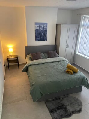 Een slaapkamer met een bed, een nachtkastje, een stadsgezicht schilderij, een garderobe en een raam met jaloezieën.