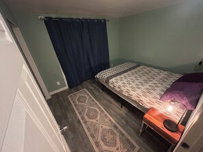 Ein Schlafzimmer mit einem Bett, einem Nachttisch, einer Lampe, einem gemusterten Teppich und einem Vorhang.