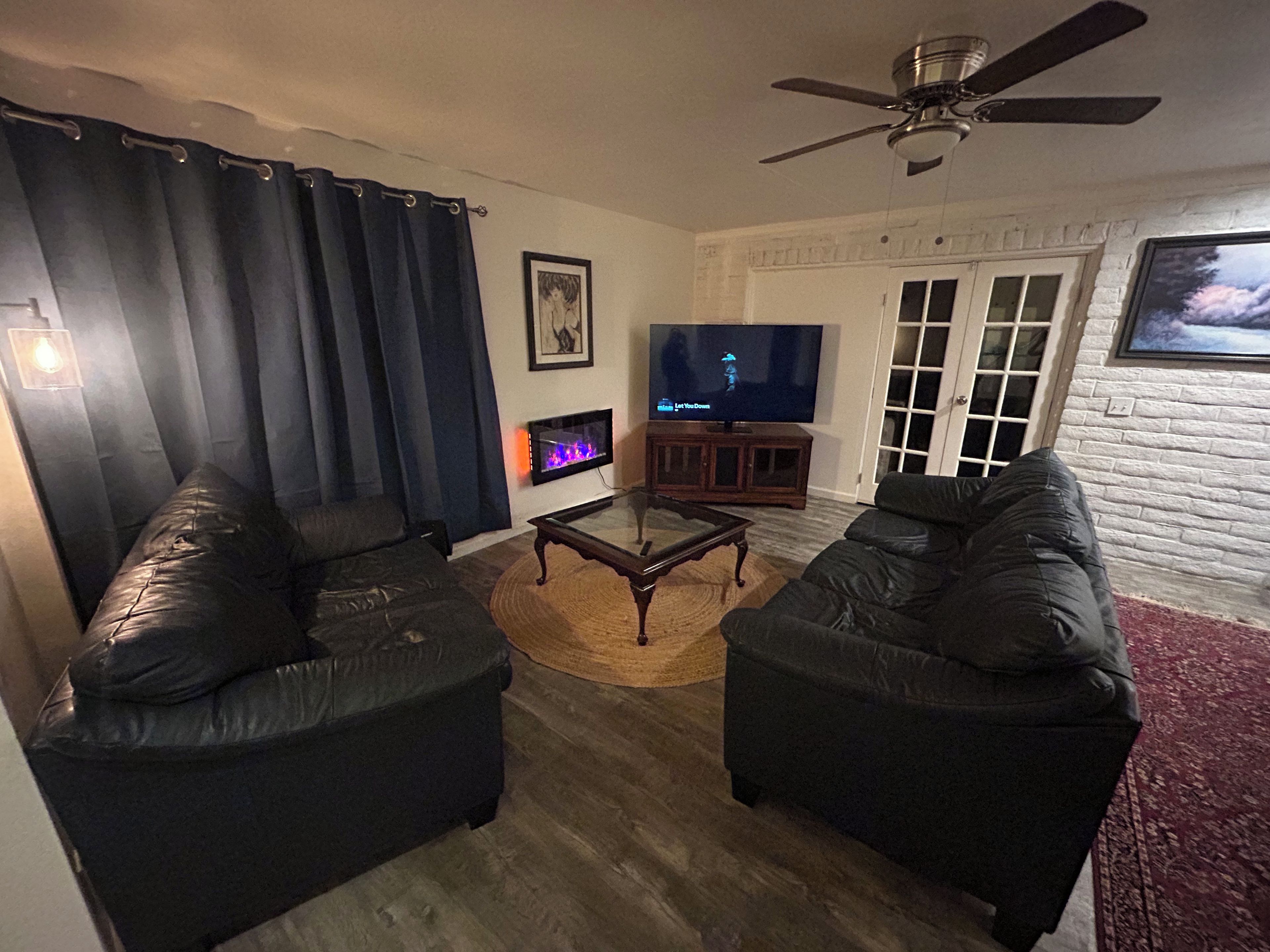 A living room with two black sofas, a glass coffee table, a fireplace, a television, and a ceiling fan.