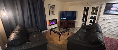A living room with two black sofas, a glass coffee table, a fireplace, a television, and a ceiling fan.