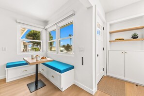 A bright kitchen with white cabinetry, a built-in bench with blue cushions, and a window overlooking a street. - Pacific Beach Bungalow - Walk to Dine, Shop and Surf (San Diego)