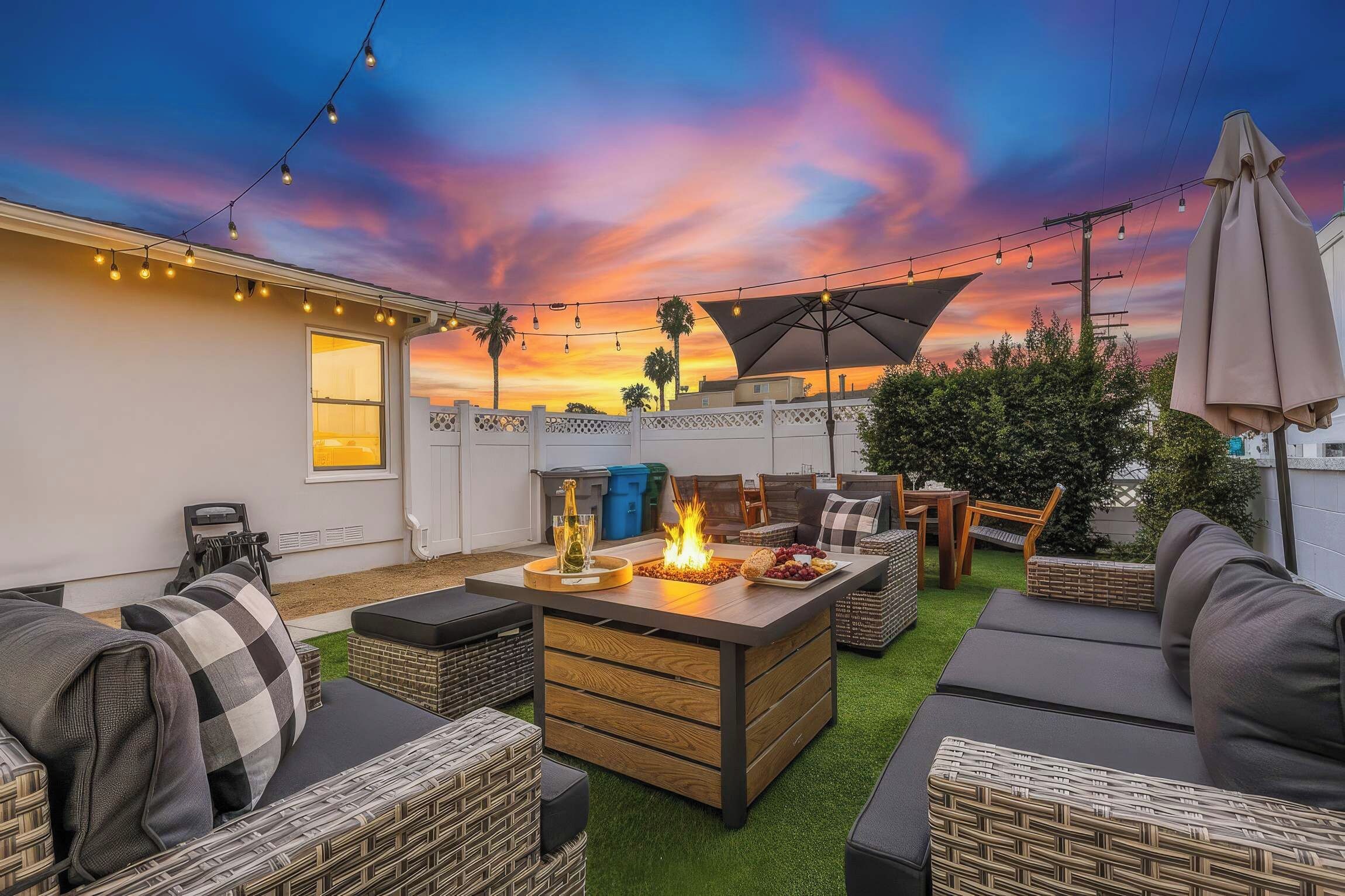 Eine Terrassengruppe im Freien mit Feuerstelle, Sitzgelegenheiten und Lichterketten vor einem Sonnenuntergangshimmel.
