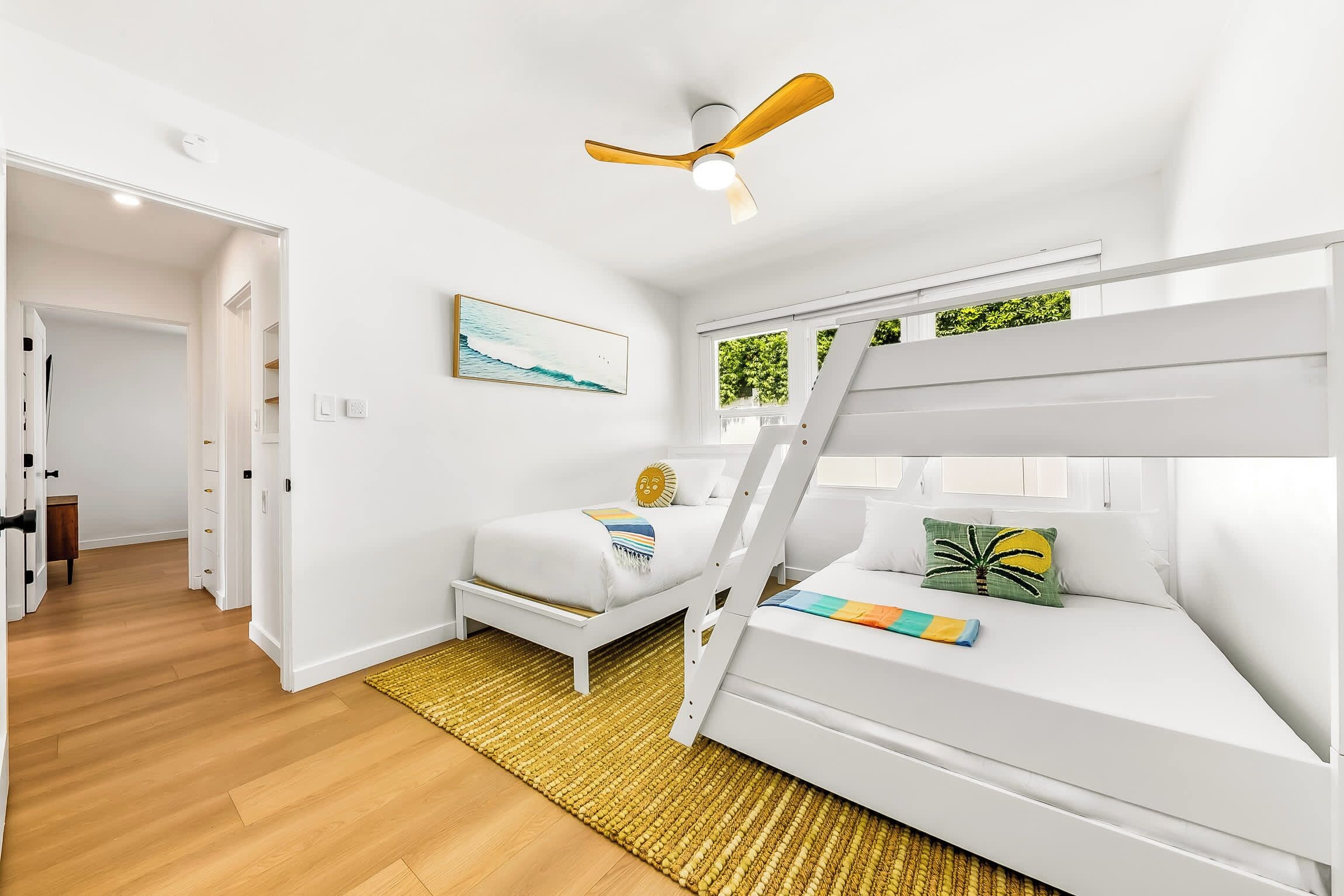 A bedroom with a bunk bed, a ceiling fan, a painting, and a view of greenery outside.