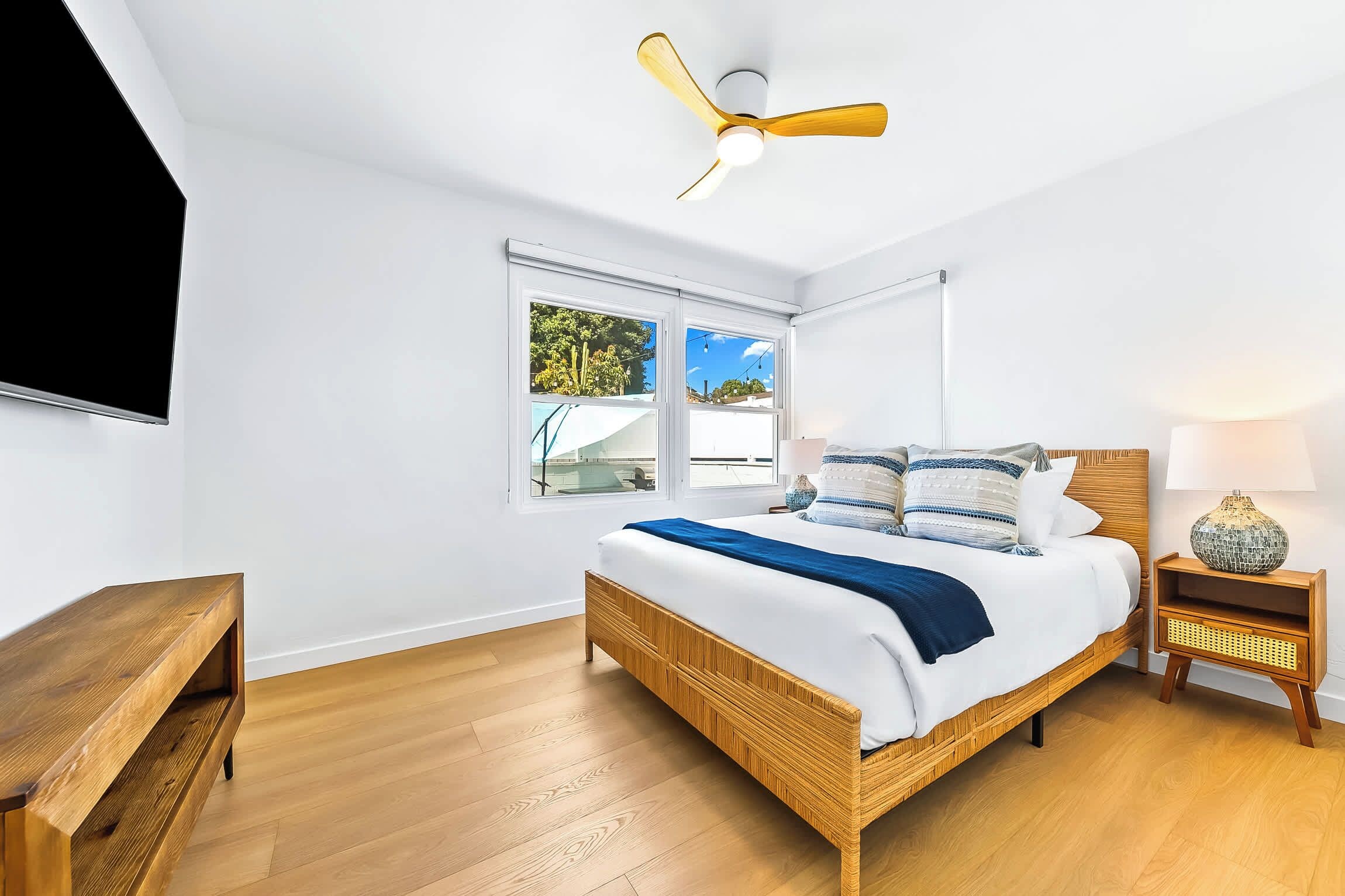 A bedroom with a bed, a nightstand, a lamp, a ceiling fan, and a window with a view of greenery.