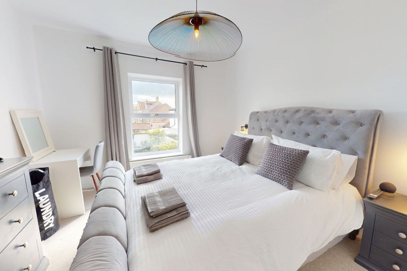 Une chambre à coucher avec un grand lit, une tête de lit rembourrée de couleur grise, une table de chevet avec une lampe et une fenêtre avec des rideaux.
