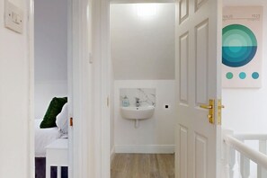 Una camera d'albergo con un letto, un cuscino verde, un muro bianco, una porta bianca, un lavandino bianco e un quadro appeso al muro.