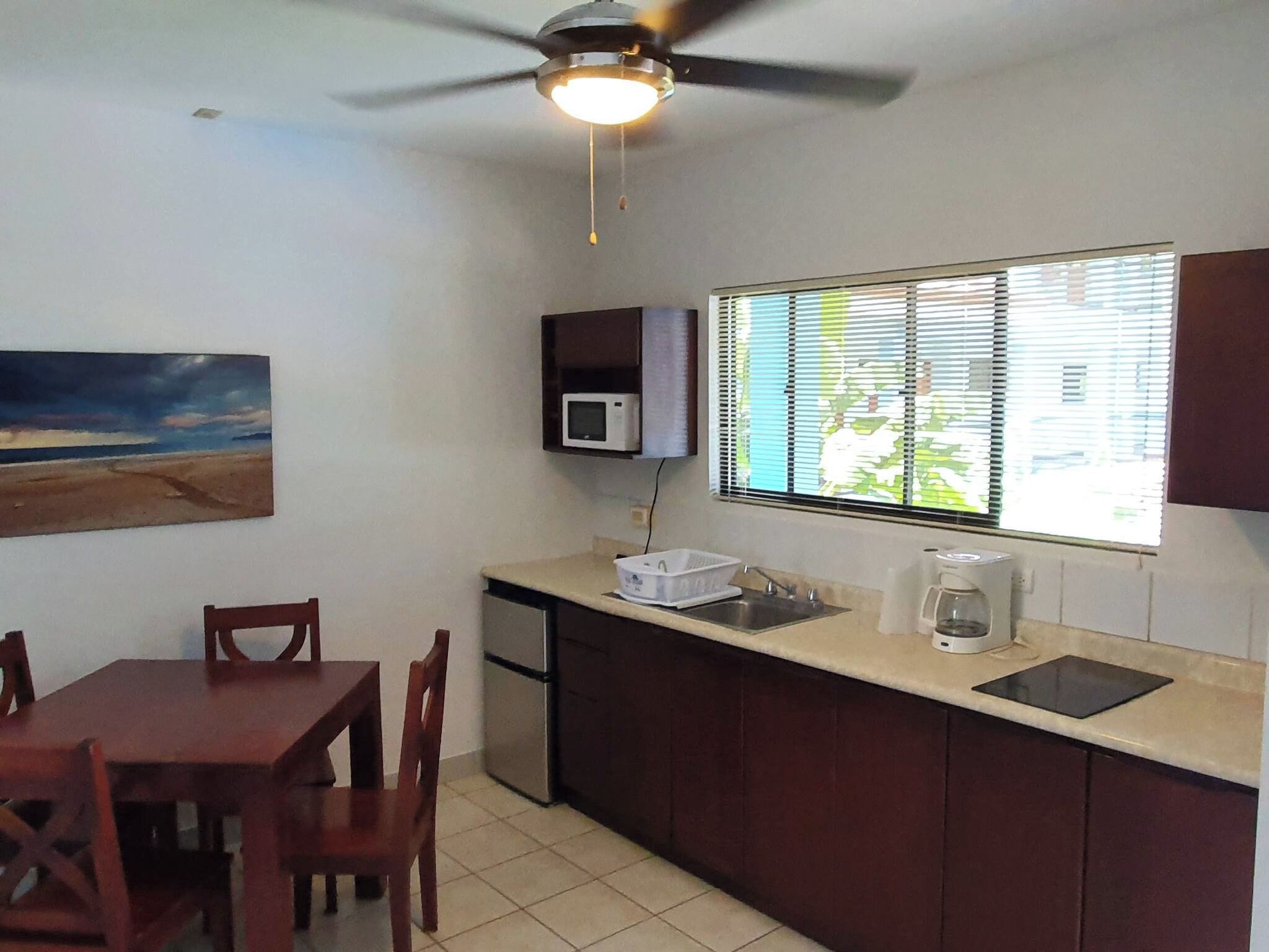 Una cocina compacta con comedor, ventilador de techo, microondas, ventana con persianas y un cuadro en la pared.