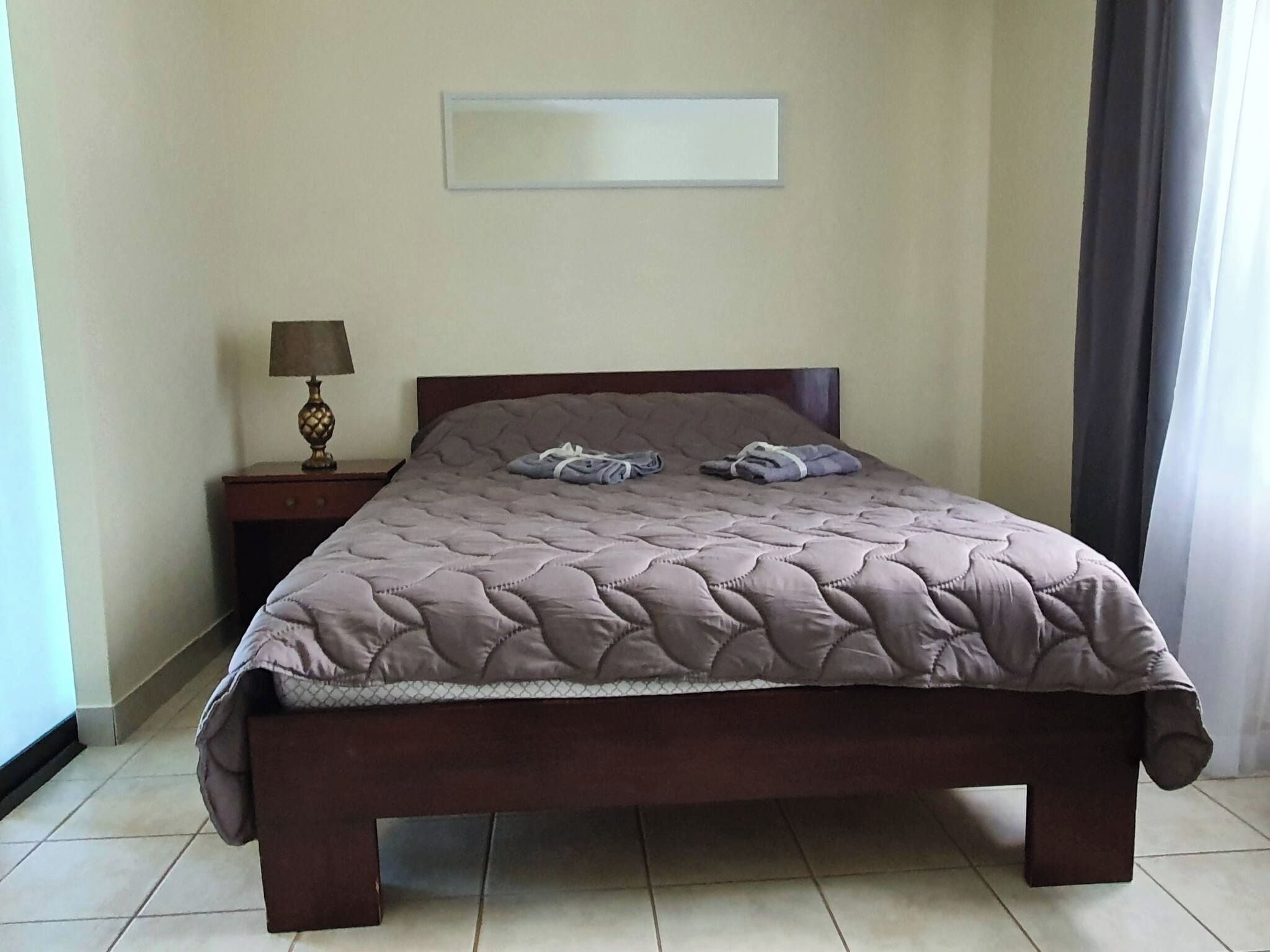 Una cama bien hecha con cubrecama acolchado, una mesita de noche con lámpara y una pared lisa con un cuadro enmarcado encima.