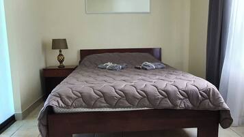 Una cama bien hecha con cubrecama acolchado, una mesita de noche con lámpara y una pared lisa con un cuadro enmarcado encima.
