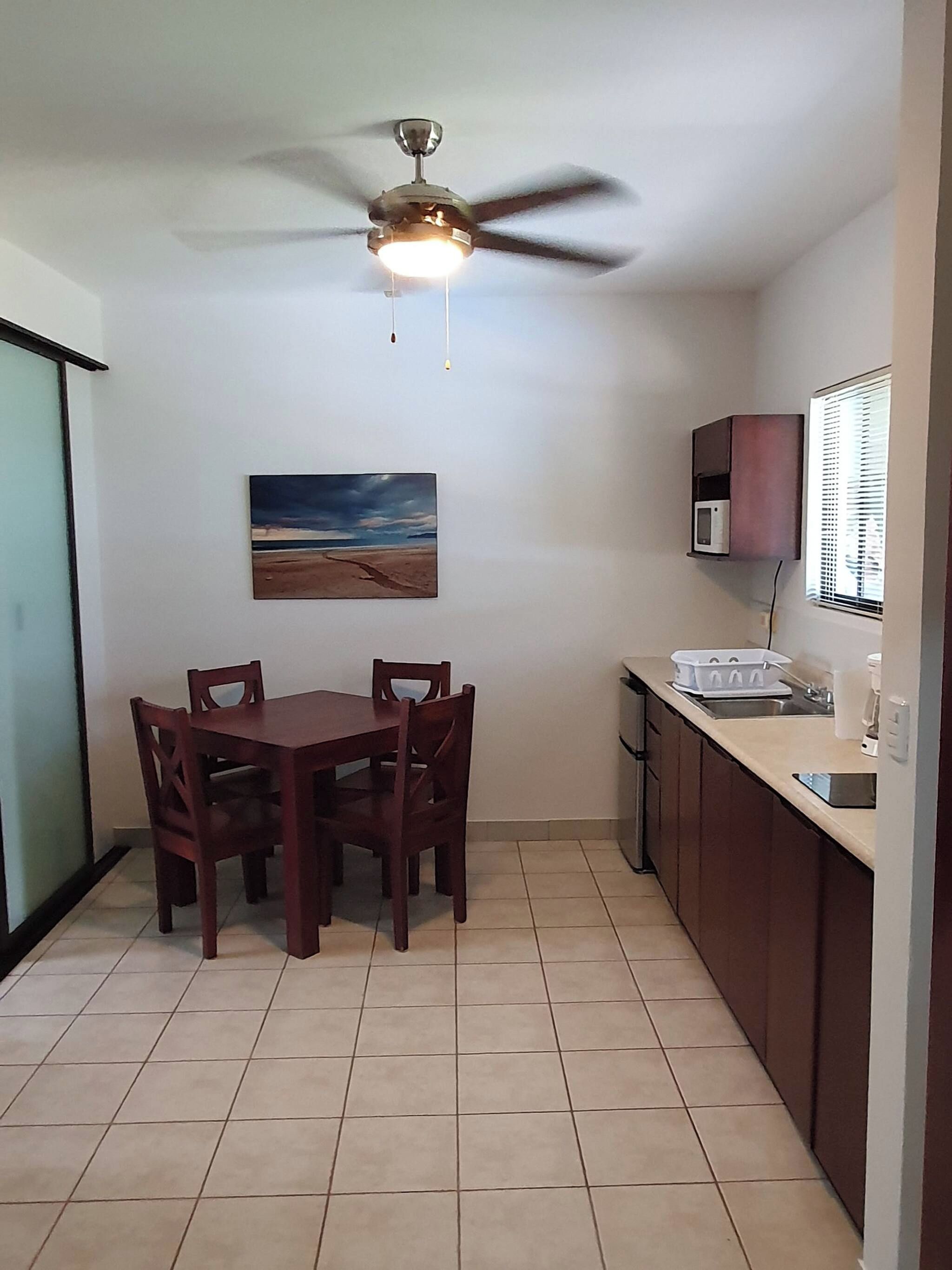 A compact kitchen with a dining table, chairs, a ceiling fan, a microwave, and a window with blinds.