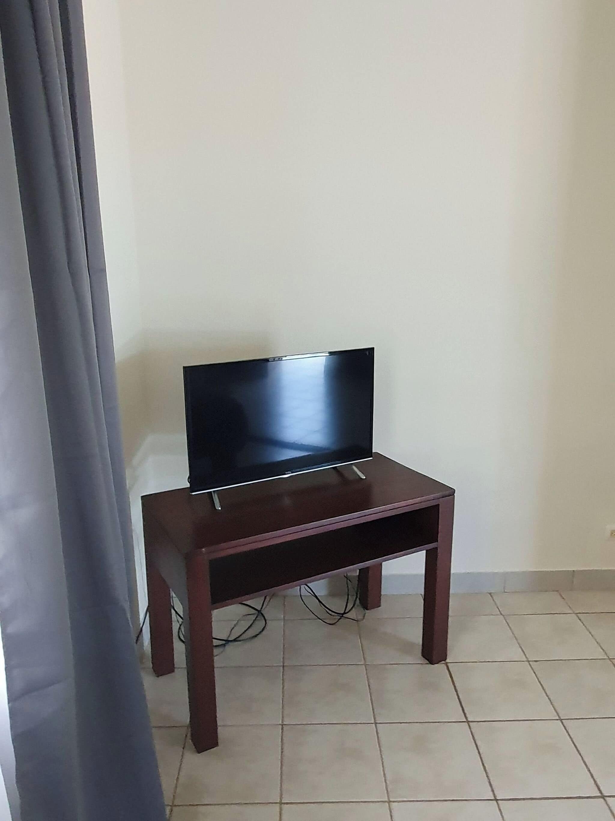 Een televisie op een houten standaard, aangesloten met een kabel, staat tegen een muur waar een gordijn gedeeltelijk zichtbaar is.