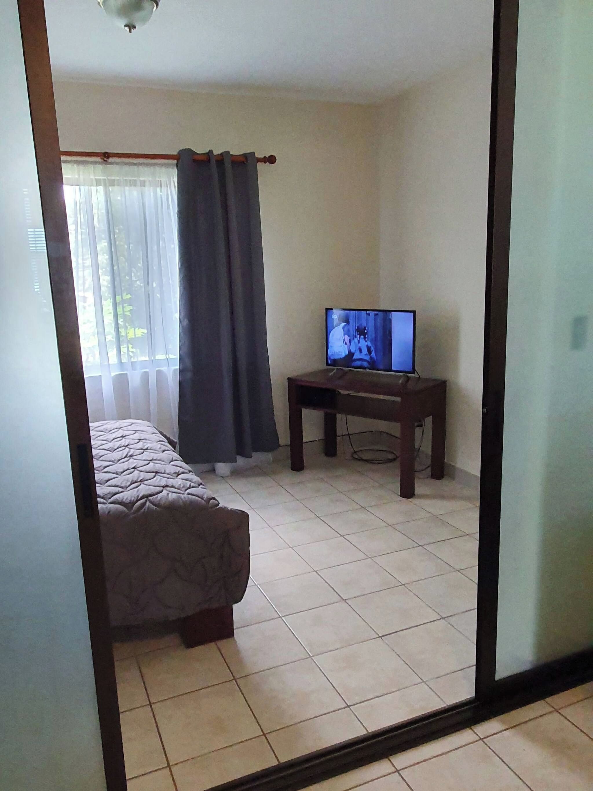 Una habitación con una cama, un televisor sobre un mueble bajo y una ventana con cortinas.