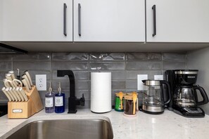 A kitchen with a stainless steel sink, a knife block, dish soap, a paper towel holder, a coffee maker, and a glass carafe. - NoDa Studio | Free Parking + Gym | Near Uptown (Charlotte)