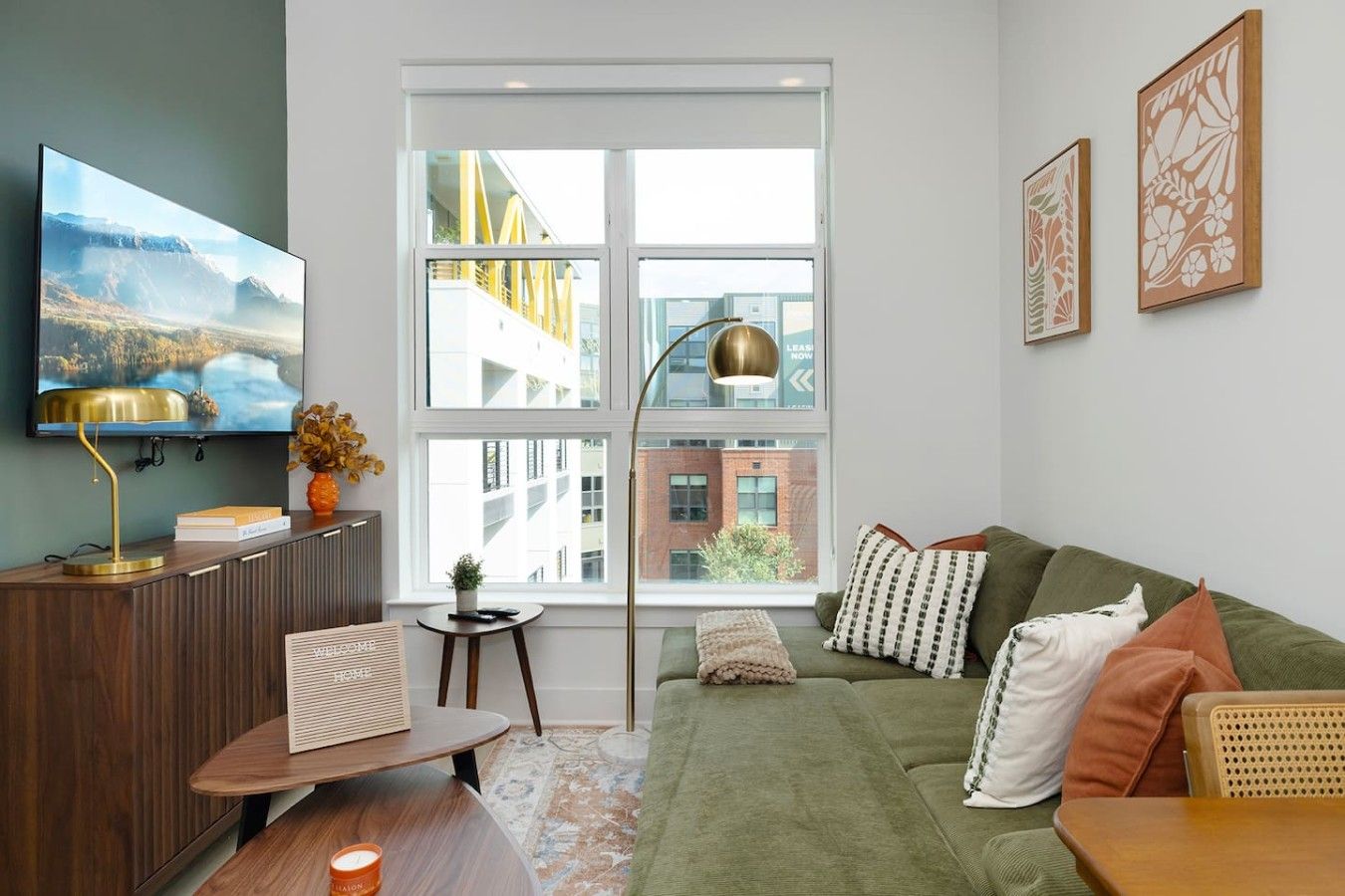 A modern living room with a green sofa, a flat-screen TV, a wooden side table, and a large window with a view of buildings.