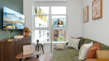 A modern living room with a green sofa, a flat-screen TV, a wooden side table, and a large window with a view of buildings.