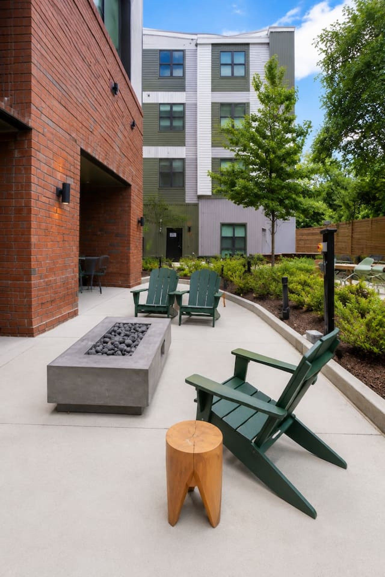 An outdoor seating area with Adirondack chairs and a fire pit, surrounded by a brick building and a modern apartment complex.