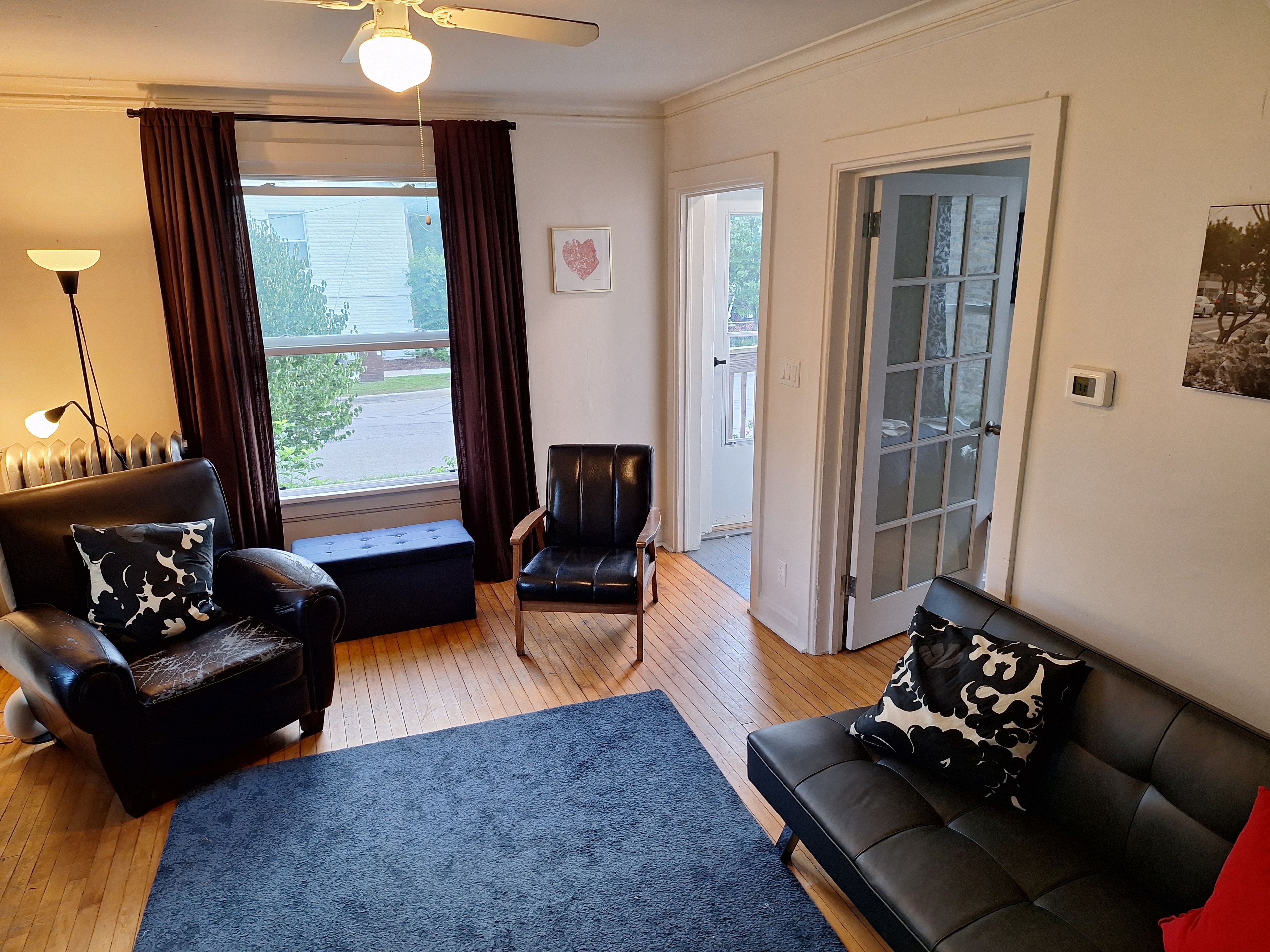 A living room with a black leather sofa, a black leather armchair, a blue rug, a floor lamp, and a window with curtains.