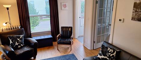 A living room with a black leather sofa, a black leather armchair, a blue rug, a floor lamp, and a window with curtains.