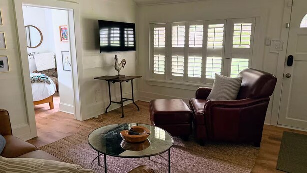 Una sala de estar con una mesa de centro de vidrio, un sillón de cuero, un televisor montado en la pared y una ventana con contraventanas.