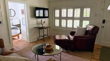 A living room with a glass coffee table, a leather armchair, a television mounted on the wall, and a window with shutters.
