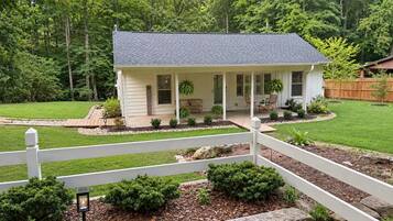 A single-story house with a porch, surrounded by a white fence, a well-maintained lawn, and a wooded area in the background.