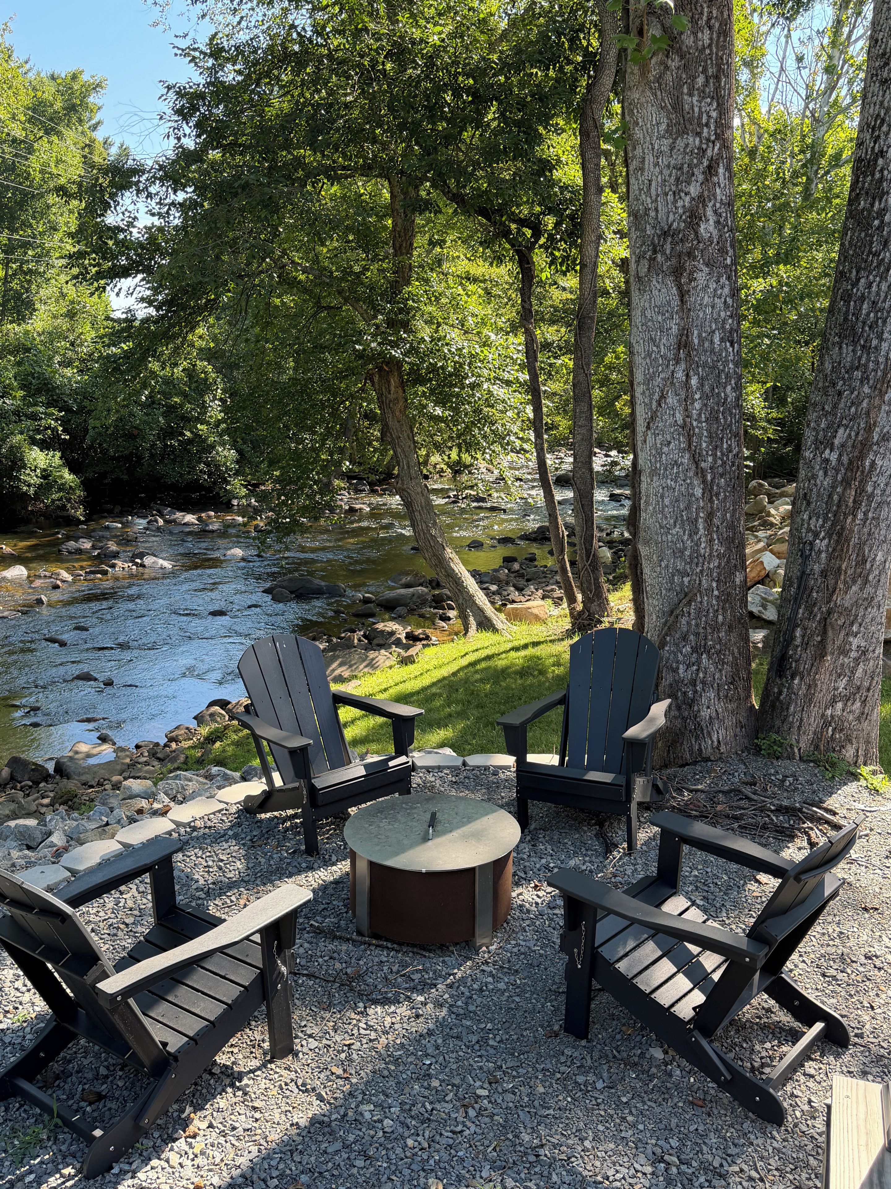 Een zithoek met Adirondack-stoelen en een ronde tafel, omringd door bomen en een beekje.