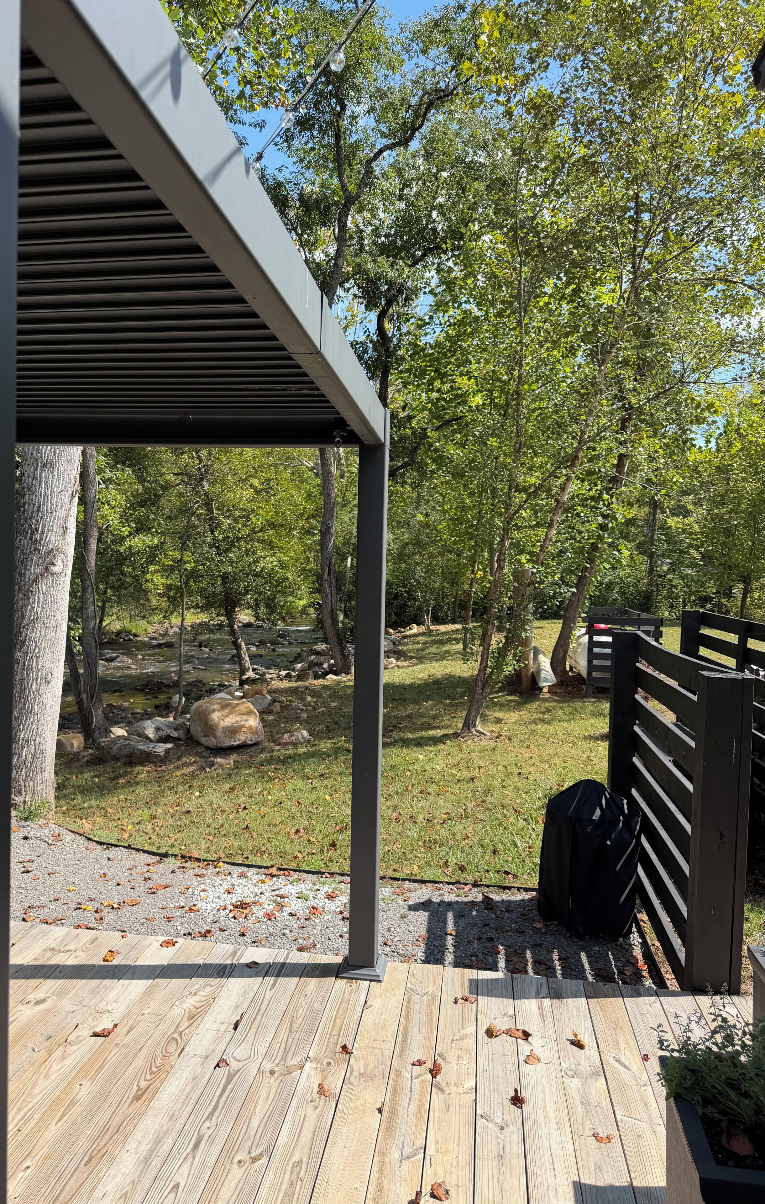 A wooden deck with a pergola structure, a fenced area with trees, and a small stream in the background.