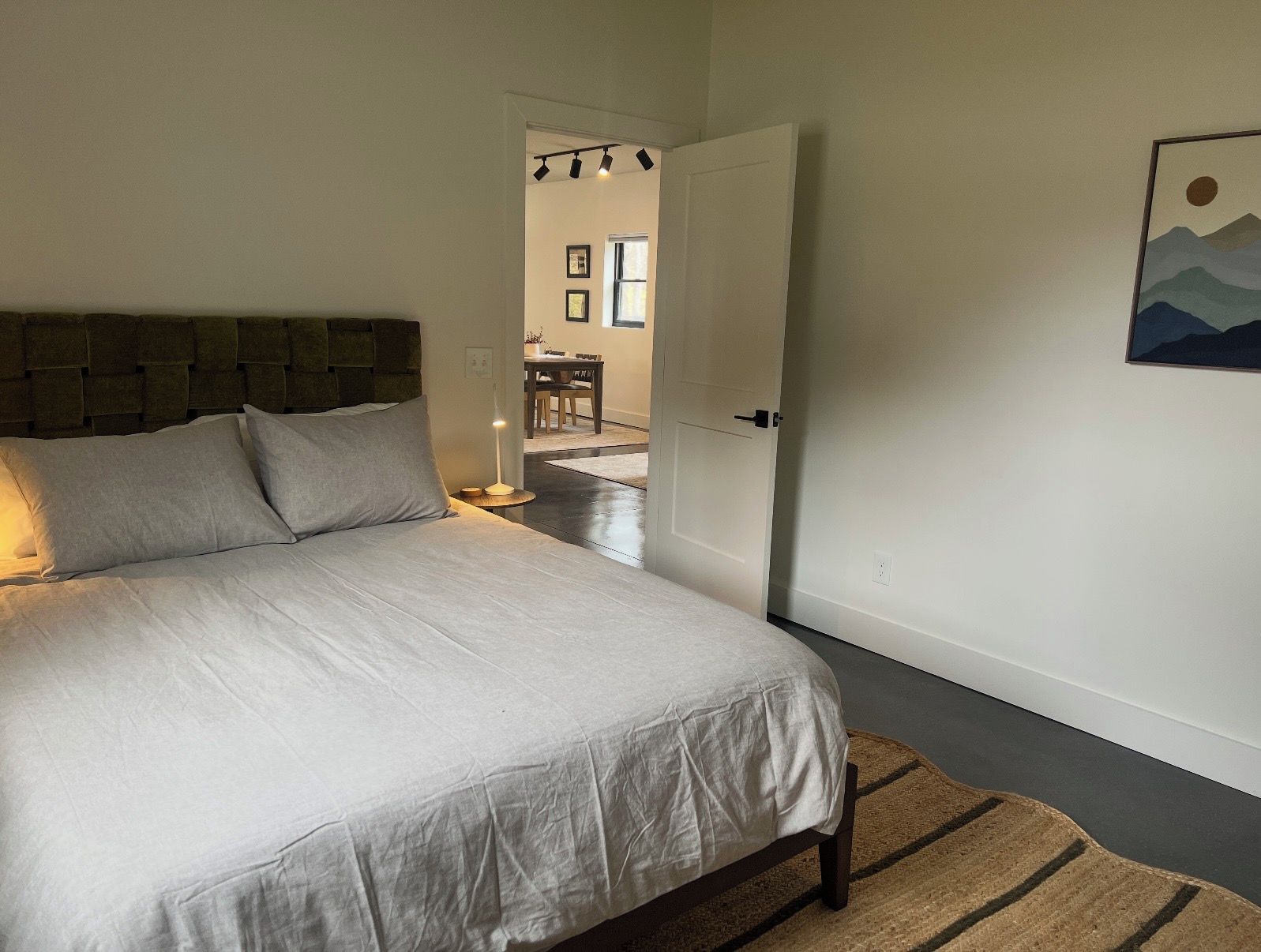 A bedroom with a bed, a nightstand, a lamp, a door leading to another room, and a framed picture on the wall.