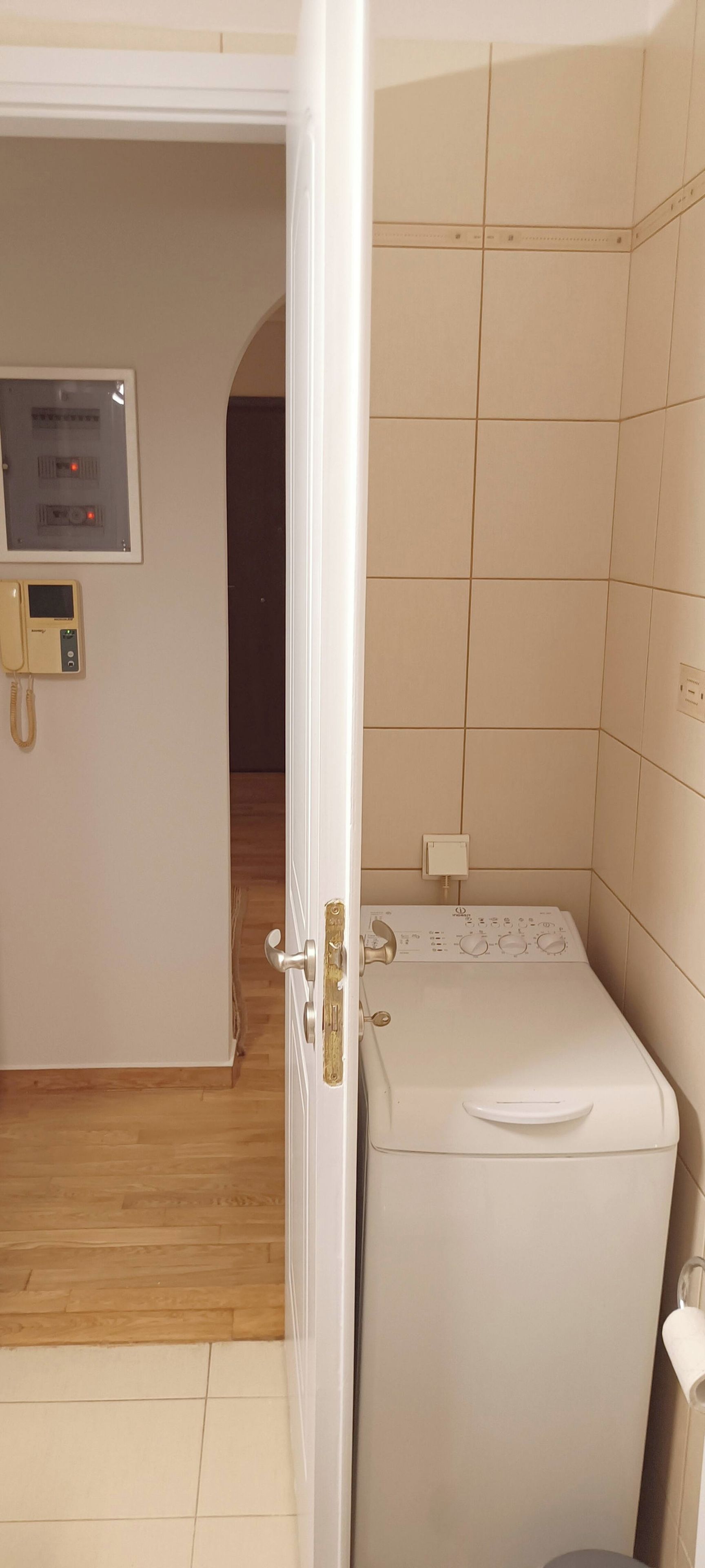 A room with a white washing machine, a wall-mounted electrical panel, and a door leading to another room.