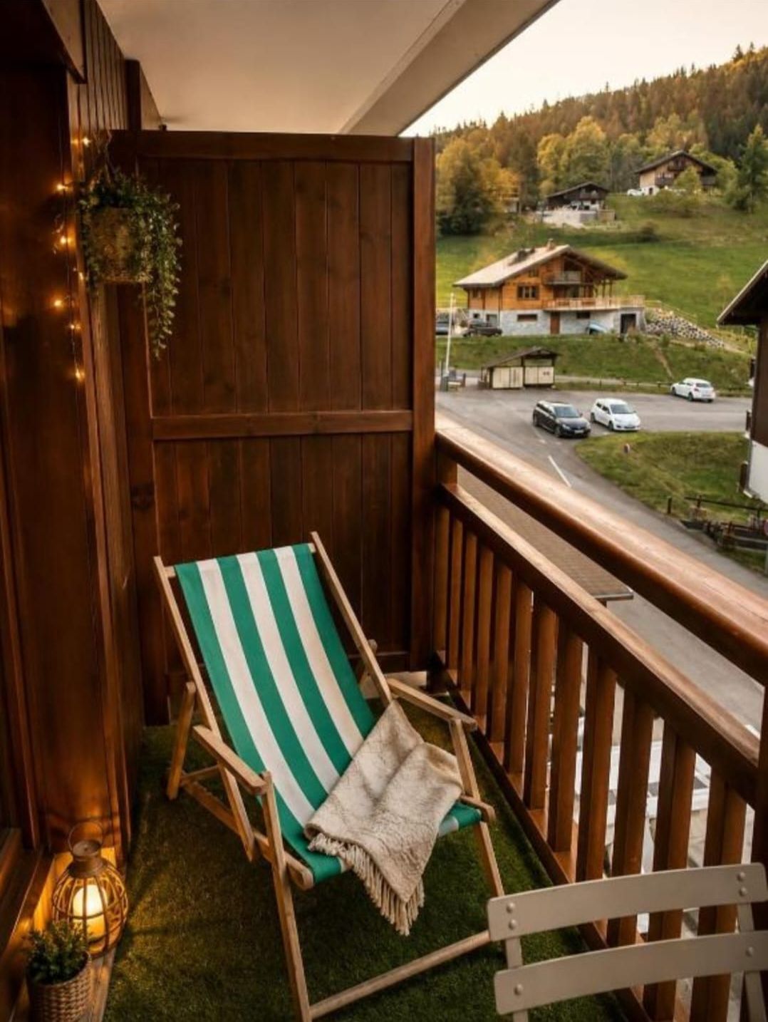 Un balcon doté d’une balustrade en bois, d’un fauteuil de jardin à rayures vertes et blanches, et d’une vue sur une route et des bâtiments au loin.