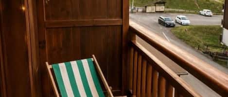 A balcony with a wooden railing, a green and white striped lounge chair, and a view of a road and buildings in the distance.