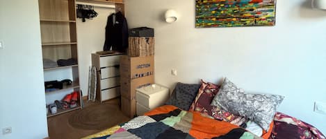 Un dormitorio con una cama, una colcha colorida, una obra de arte en la pared y un armario con ropa colgada.
