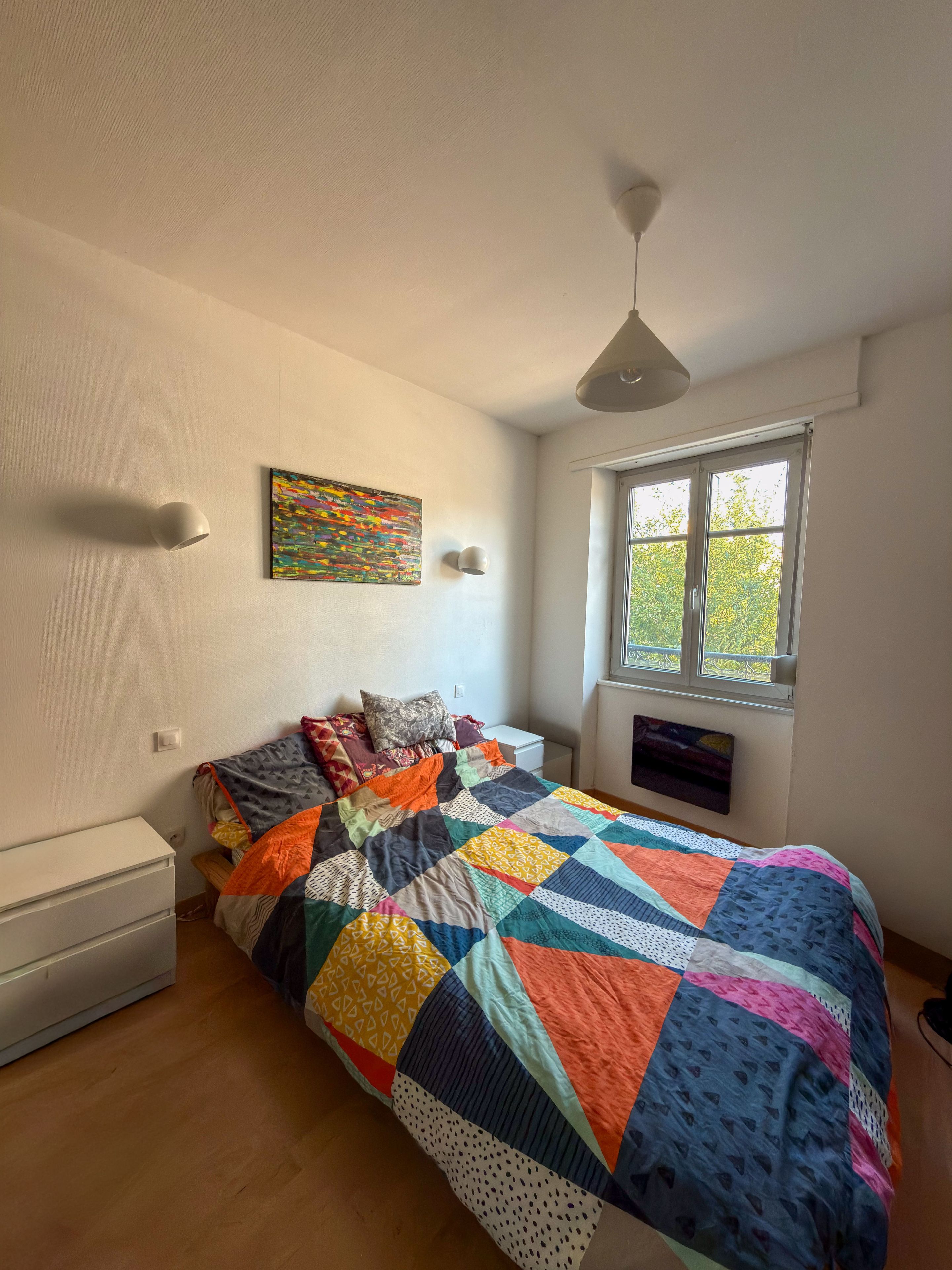 A bedroom with a bed, a window, a wall-mounted artwork, and a ceiling light.