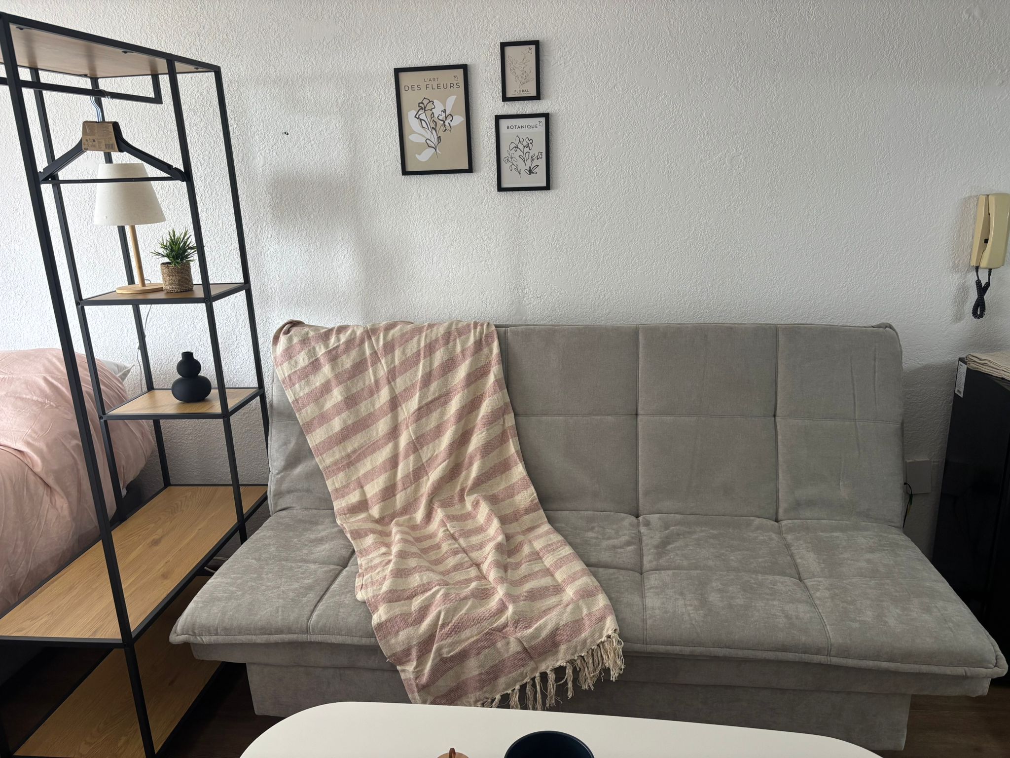 Uma sala de estar com um sofá cinza, um cobertor listrado, uma estante de metal com uma luminária, e duas gravuras botânicas emolduradas na parede.