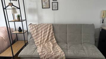 Uma sala de estar com um sofá cinza, um cobertor listrado, uma estante de metal com uma luminária, e duas gravuras botânicas emolduradas na parede.