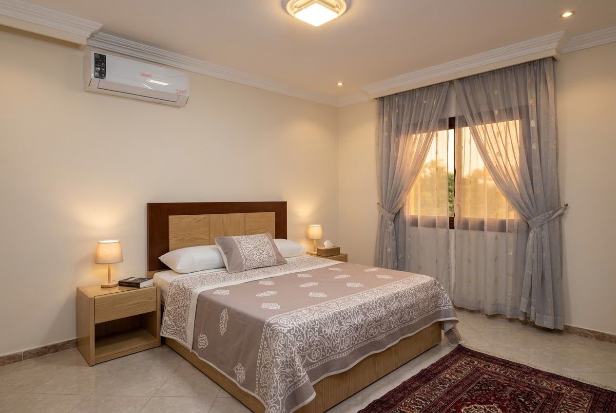 A hotel room with a bed, bedside lamps, an air conditioning unit, a window with curtains, and a rug.