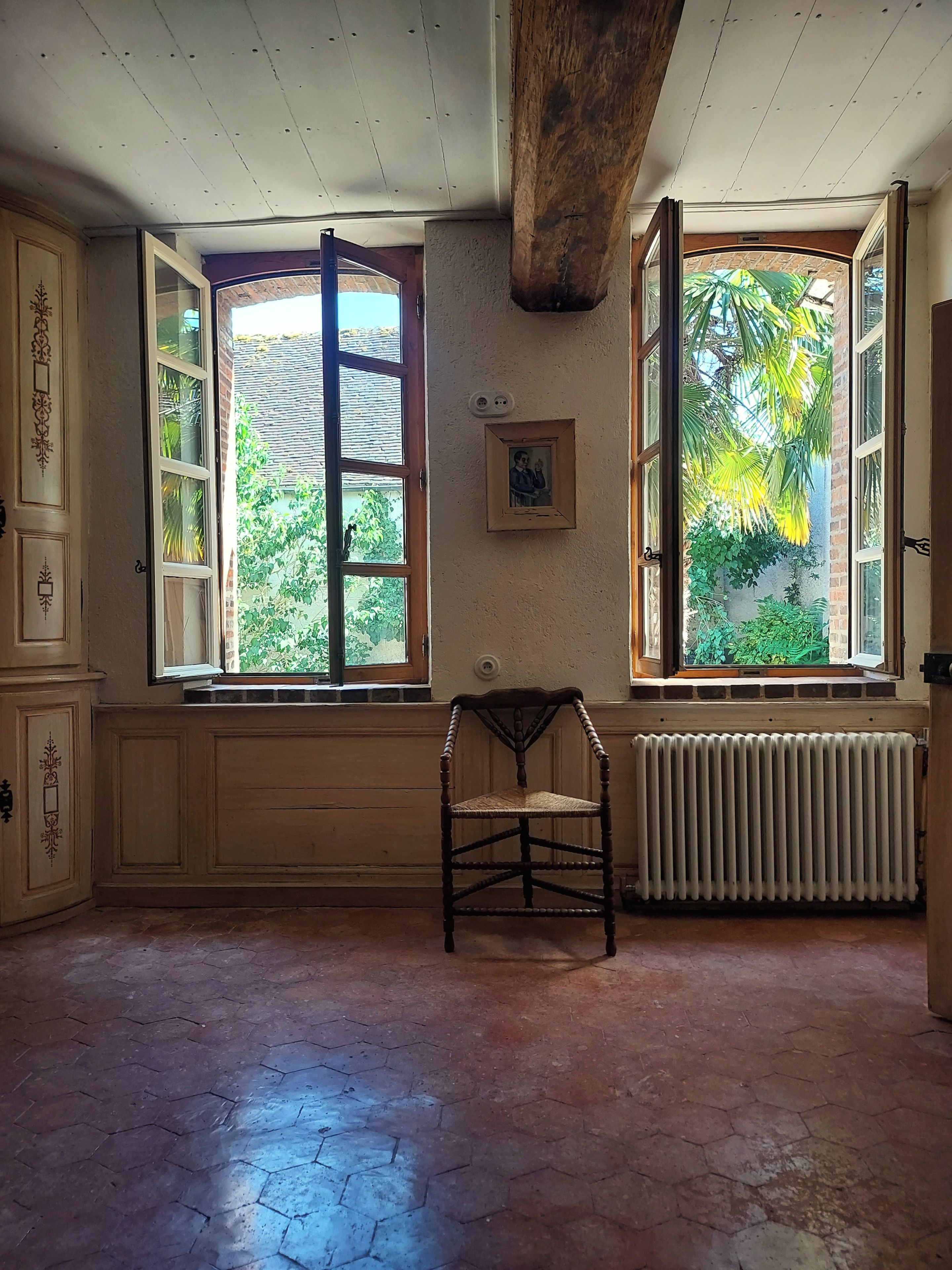 A room with a wooden chair, a radiator, and two open windows with a view of greenery outside.