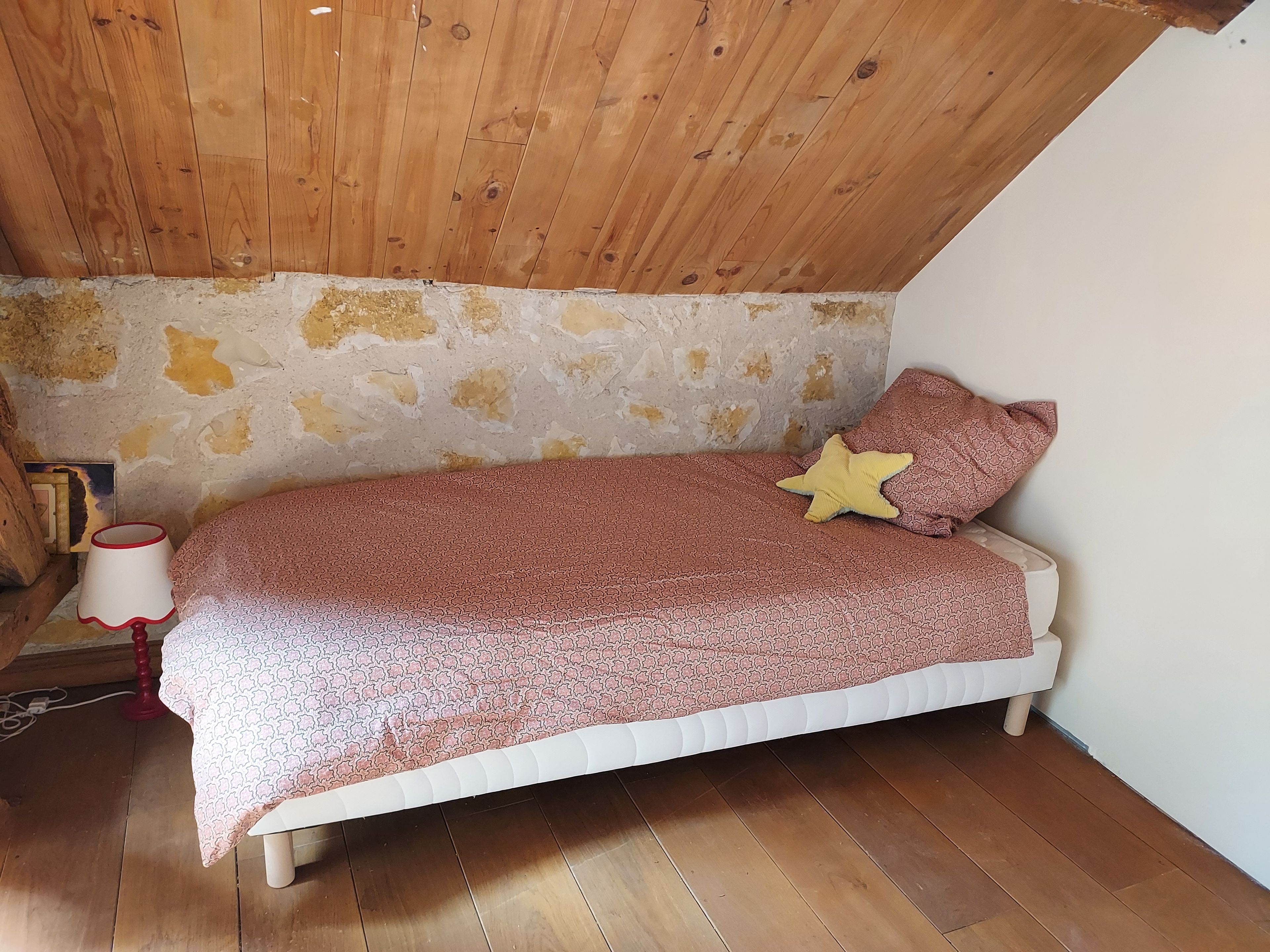 Una habitación pequeña y acogedora con techo inclinado, una cama individual con ropa de cama estampada, un cojín con forma de estrella y una lámpara sobre suelo de madera.