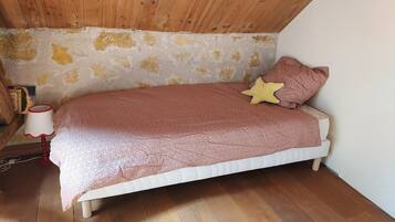 Una habitación pequeña y acogedora con techo inclinado, una cama individual con ropa de cama estampada, un cojín con forma de estrella y una lámpara sobre suelo de madera.