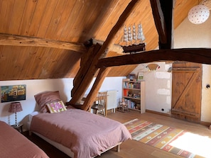 Quarto de s ótão acolhedor com vigas de madeira aparentes, cama com manta rosa, estante de livros e um pequeno modelo de navio.