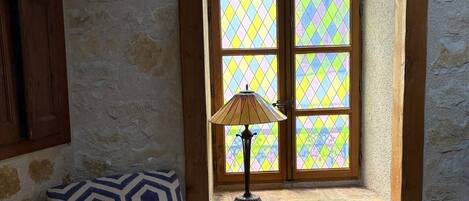 Ein Zimmer mit einem gemusterten Sessel, einem Fenster mit Buntglas und einer Lampe auf der Fensterbank.