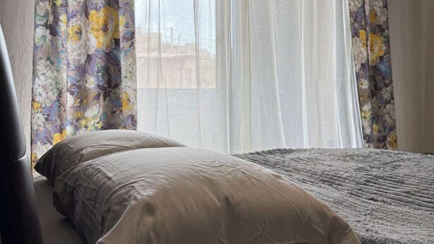 A room with a bed, a window with sheer curtains, and a view of the outside. - Monte Cara (Mindelo)