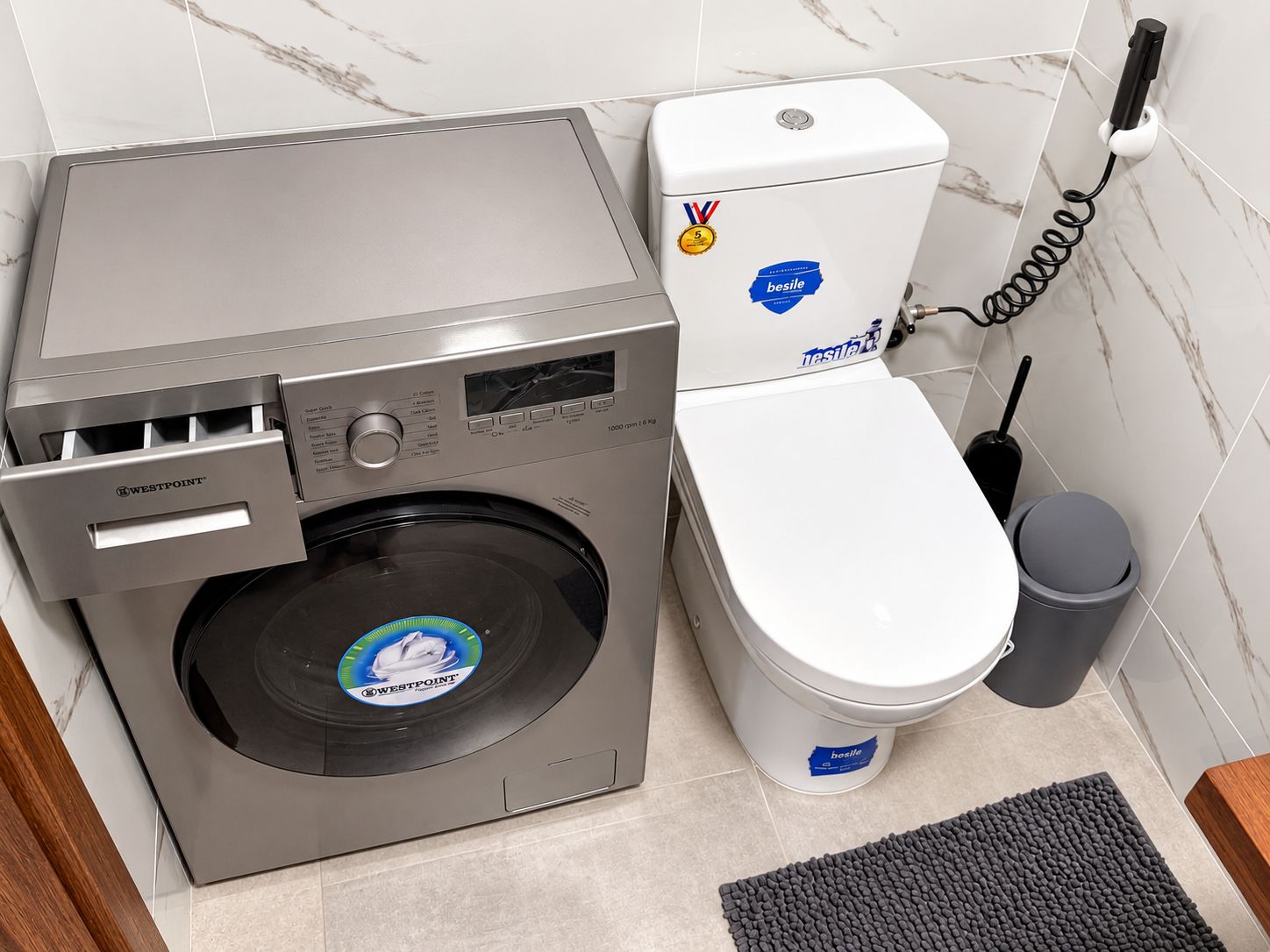 A compact bathroom with a WestPoint washing machine, a toilet, and a trash bin.