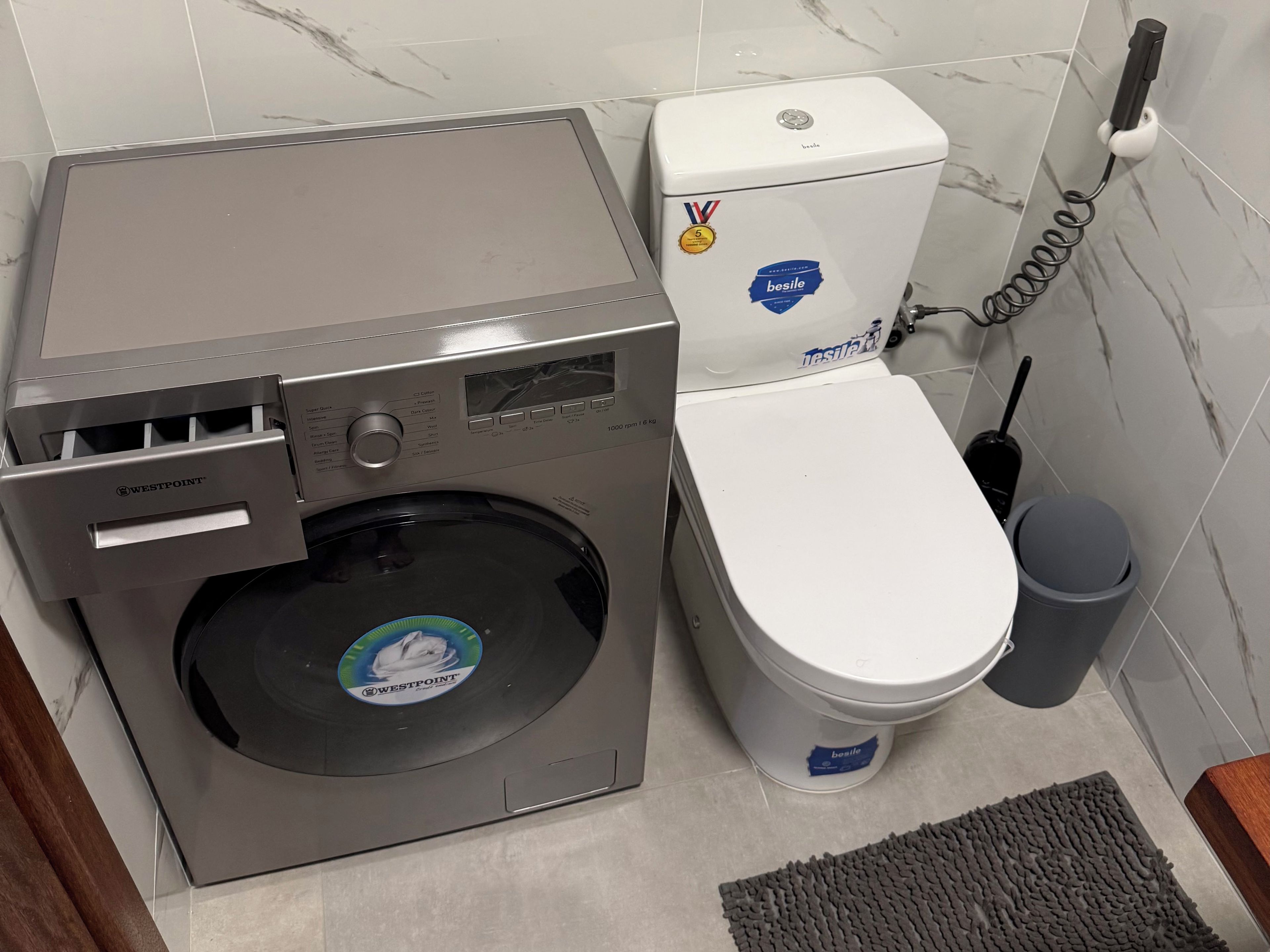 A compact washer and dryer unit with a control panel, a toilet with a bidet attachment, and a trash bin.