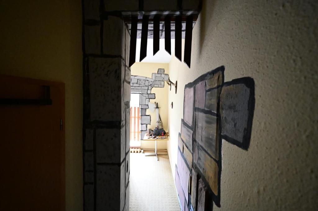 A narrow hallway with a wooden door on the left, a stone wall with a brick pattern on the right, and a staircase leading to an upper level.